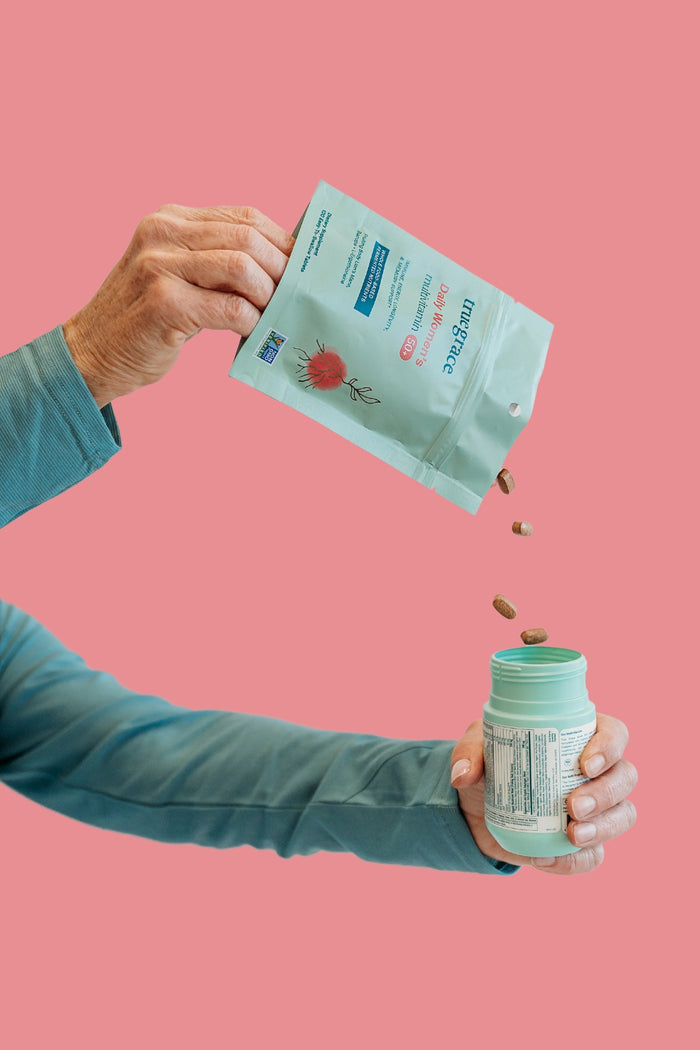 A hand pours supplements from a blue-green pouch adorned with a floral design and labeled Daily Women’s 50+ Multivitamin by True Grace into a jar against a pink background, capturing the essence of health and wellness.