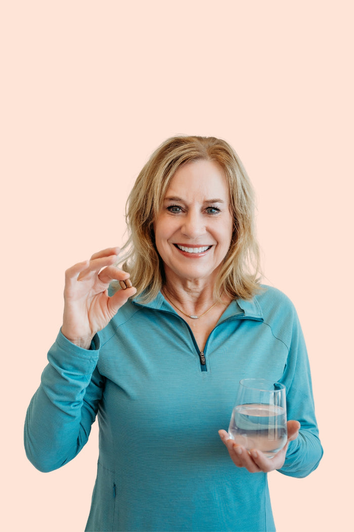 A woman in a blue top smiles, holding a glass of water in one hand and True Grace Daily Women’s 50+ Multivitamin in the other against a beige background.