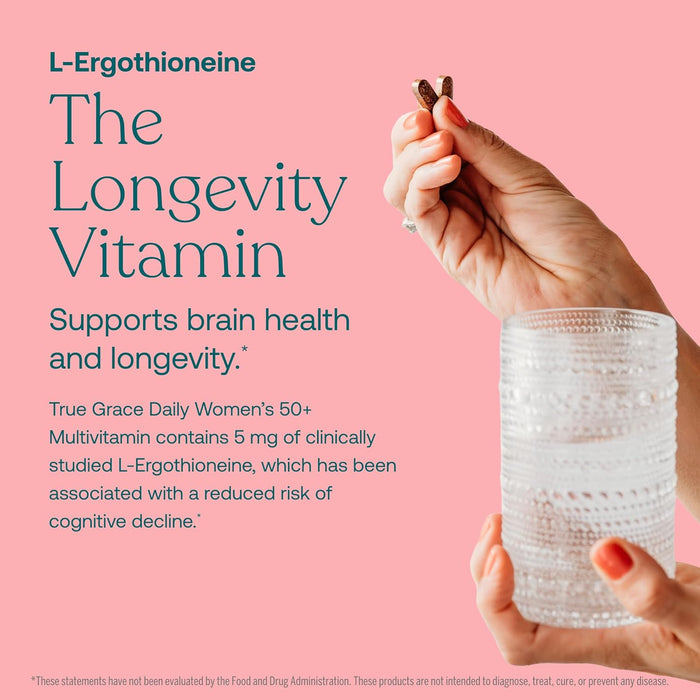 A hand holds two brown capsules above a glass of water on a pink background. True Grace Daily Women’s 50+ Multivitamin Tablets offer longevity and memory support, specially designed for women aged 50+.