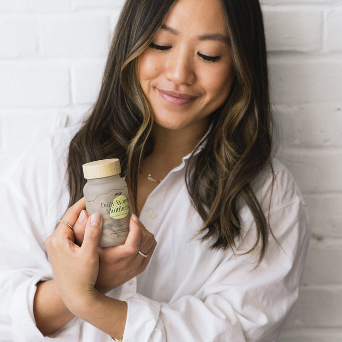 A person with long hair and a white shirt smiles while holding a bottle labeled Daily Womens Multiherbal - For Total Body + Cycle Balance by WTHN, against a white brick wall.