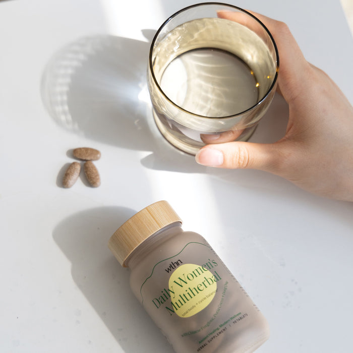 A hand holds a glass of water on a white surface next to a wooden-capped bottle labeled Daily Womens Multiherbal - For Total Body + Cycle Balance by WTHN. Three oval tablets nearby cast soft shadows in the sunlight, promoting hormonal balance.
