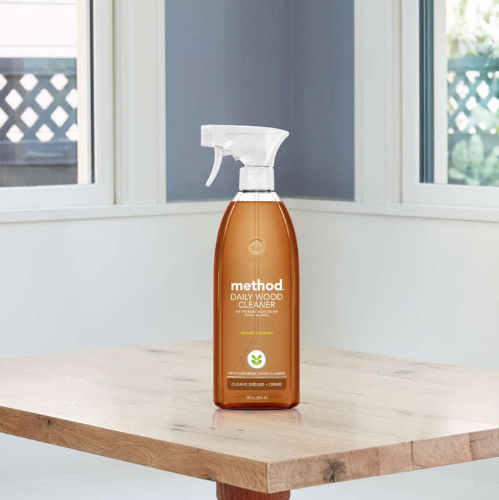 A bottle of method Daily Wood Cleaner with an almond scent sits on a wooden table in a bright room with large windows. The cruelty-free cleaner, packaged in recycled plastic, has a transparent body and white spray nozzle.