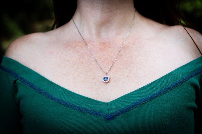 A close-up of a person's neck and chest wearing a delicate silver chain necklace with a small heart-shaped cremation silver pendant featuring a blue center stone.