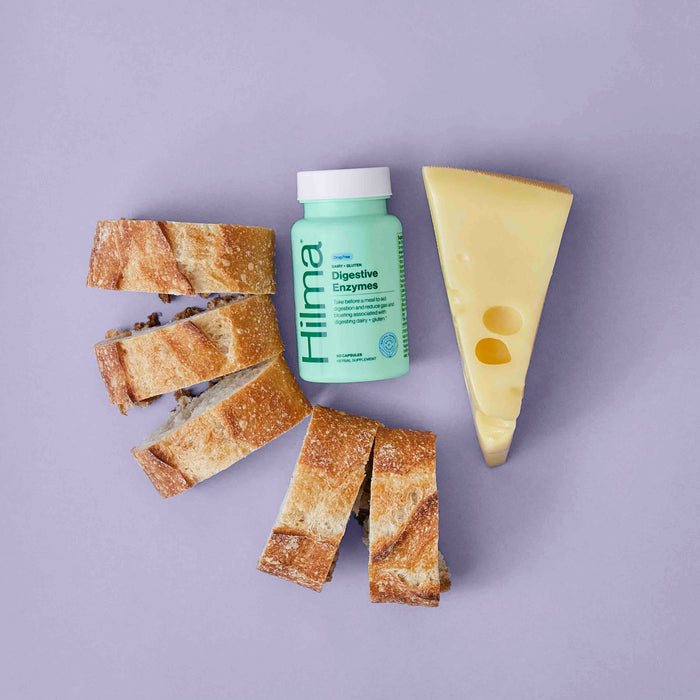 A green bottle of Hilmas Dairy + Gluten Digestive Enzymes is nestled between three slices of crusty bread and a wedge of Swiss cheese with two holes, set against a light purple background—ideal for promoting optimal dairy and gluten digestion.