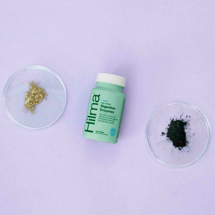 A green bottle labeled Hilma Dairy + Gluten Digestive Enzymes is centered on a light purple background, flanked by petri dishes—one with light and the other dark green powder—emphasizing its support for gluten digestion and overall digestive health.