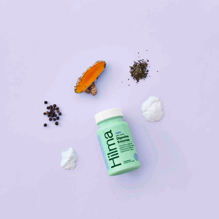A green bottle labeled Hilma Dairy + Gluten Digestive Enzymes is placed on a light purple background, surrounded by natural ingredients like turmeric, black peppercorns, herbs, and two mounds of white powder.