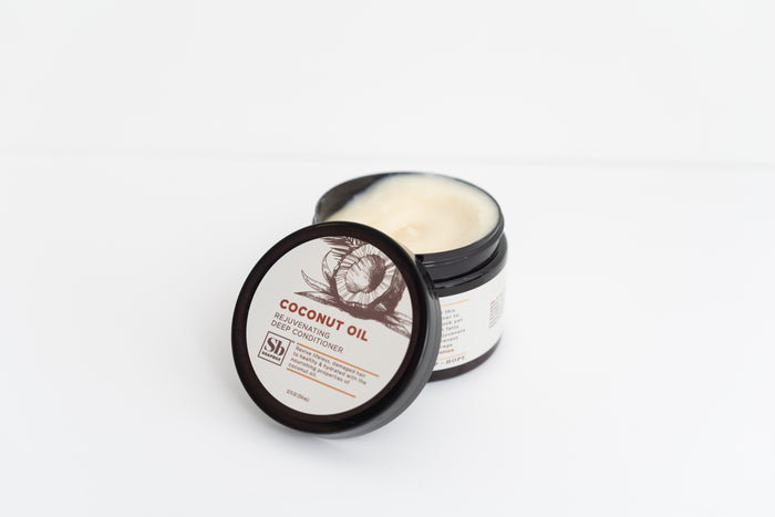 A slightly open black jar reveals Soapboxs Deep Conditioner - Moisturizing Coconut Oil Jar, displaying the creamy contents inside. The label showcases a coconut illustration and describes the sulfate-free formula, all set against a plain white background.