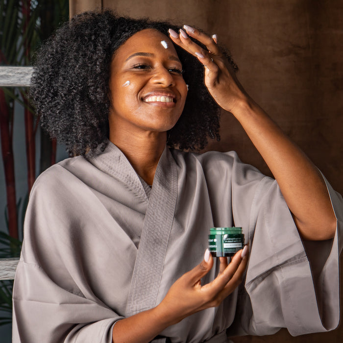 A person in a gray robe smiles while applying Tree To Tubs Deep Hydrating Moisturizer for Sensitive Skin with Green Tea to their face. Holding a small green jar, they enjoy skincare bliss against a backdrop of curtains and plants.
