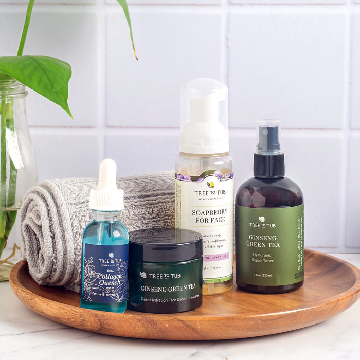 A wooden tray showcases Tree To Tub skincare items, including a Deep Hydrating Moisturizer for Sensitive Skin with Green Tea, a dropper bottle, foaming cleanser, and spray toner. A gray rolled towel and green plant enhance the calm setting against a tiled wall.
