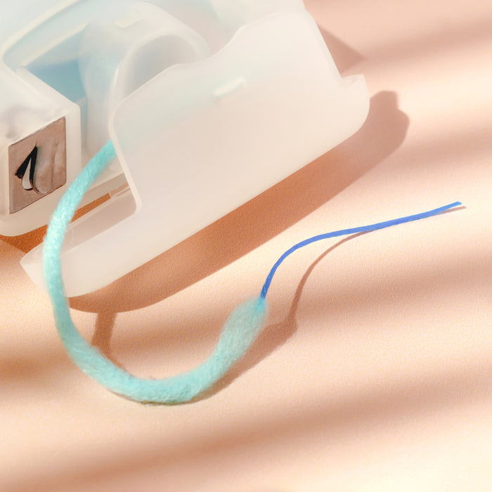 A close-up of a partially unspooled tape dispenser with a strip of blue tape attached to a Cocofloss Dental Floss + Threaders fuzzy cord. Soft, warm lighting casts gentle shadows on the peach-colored surface.