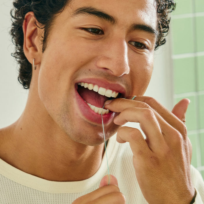 In a brightly lit bathroom with green tiles, a curly-haired person joyfully flosses their teeth using Cocoflosss gum-soothing dental floss. Theyre wearing a recycled polyester white shirt and sporting a hoop earring.