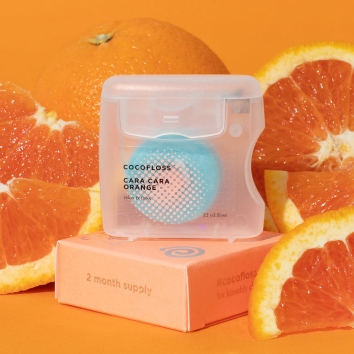 A Cocofloss dental floss package, labeled Cara Cara Orange with gum-soothing coconut oil, sits on a pink 2-month supply box. The recycled polyester plastic case is surrounded by fresh Cara Cara orange slices against an orange backdrop.