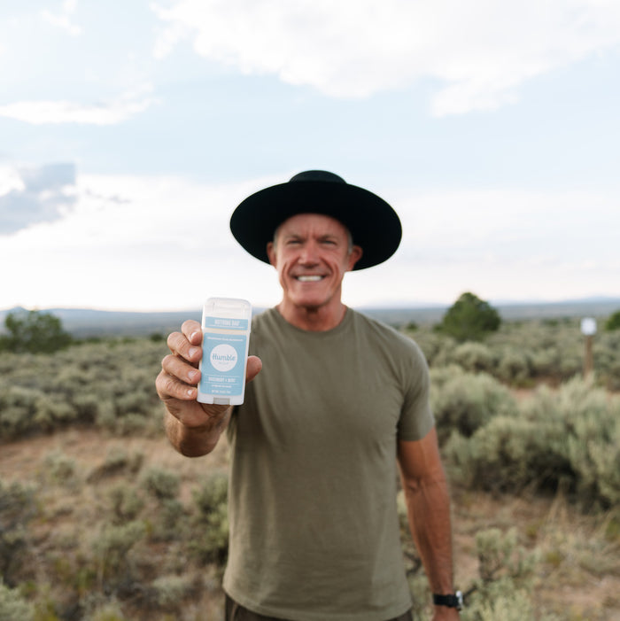 In a partly cloudy field with serene shrubs, a smiling person in a black hat and green shirt proudly holds up a Humble Brands Deodorant - Aluminum Free stick. The cruelty-free product is packaged in recycled plastic.