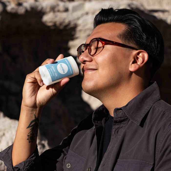 A person with glasses and a dark shirt smiles with closed eyes, holding a light blue and white Humble Brands Aluminum Free Deodorant near their face outdoors, relishing its fresh scent from the cruelty-free formula and recycled plastic packaging.