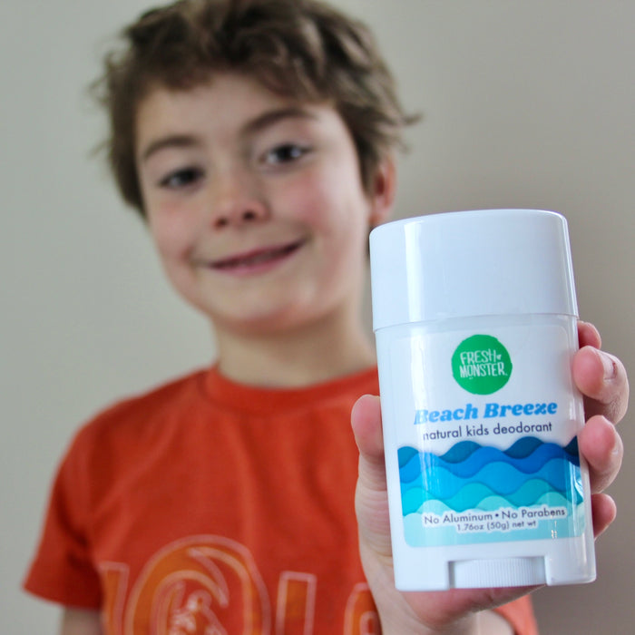 A child in an orange shirt smiles, proudly holding up a stick of Fresh Monsters Deodorant for Kids and Teens. Its green label highlights its aluminum and paraben-free formula, designed for gentle care on young, sensitive skin.