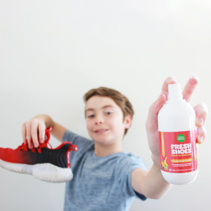 A boy in a gray shirt holds a red and black sneaker and Fresh Monsters Deodorizing Shoe Spray, which uses bio-enzymatic technology for odor elimination, against a plain white background.