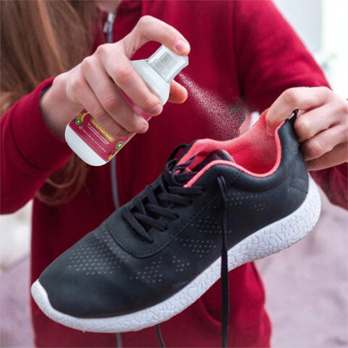 Wearing a red hoodie, someone uses Fresh Monsters Deodorizing Shoe Spray with bio-enzymatic technology to freshen their black and pink athletic shoe, holding it in one hand and operating the spray bottle with the other for a naturally fresh scent.