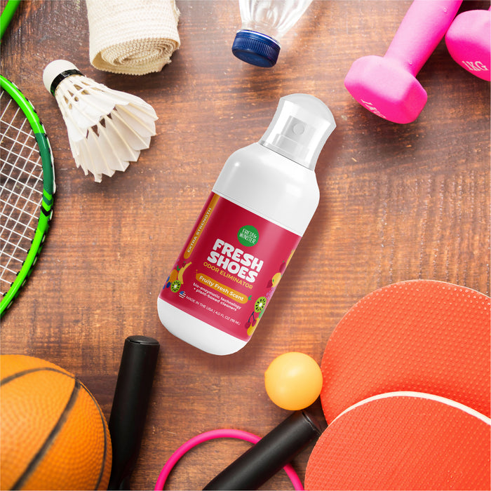A Fresh Monster Deodorizing Shoe Spray with bio-enzymatic technology rests on a wooden surface, surrounded by sports equipment like a shuttlecock, basketball, ping pong paddles and ball, tennis racket, pink dumbbells, and a white sports towel.