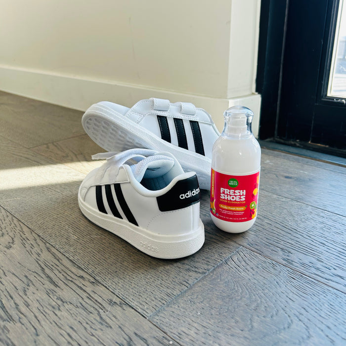 White sneakers with black stripes rest on a wooden floor next to a bottle of Fresh Monsters Deodorizing Shoe Spray, famous for its natural fragrance. Sunlight streams in through a nearby glass door, emphasizing the fresh allure of both the shoes and spray.