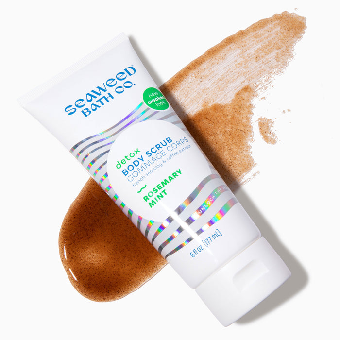 A white tube of The Seaweed Bath Cos Detox Exfoliating Body Scrub in Rosemary Mint flavor rests on a smear of brown scrub, featuring a green and blue holographic design. It highlights natural ingredients like bladderwrack seaweed and French clay for exceptional exfoliating benefits.
