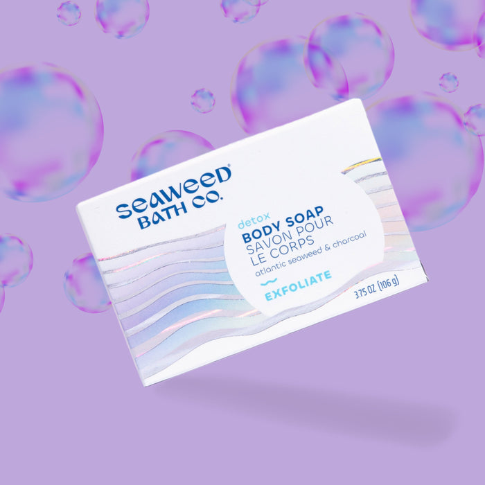 The Seaweed Bath Cos Detox Exfoliating Body Soap blends Atlantic and bladderwrack seaweed extract with charcoal, shown against a purple background with translucent bubbles. Its packaging features light wave patterns.