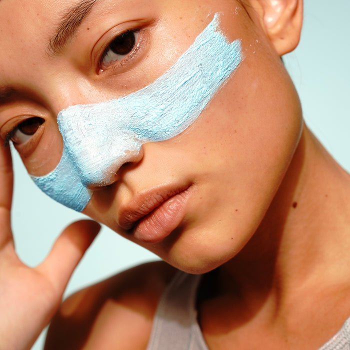 A person with light skin poses close to the camera, wearing a thick band of FRÉ Skincare's DETOX ME Instant Clearing Mask with natural kaolin clay across their nose and under both eyes, touching their temple and looking directly at the camera.