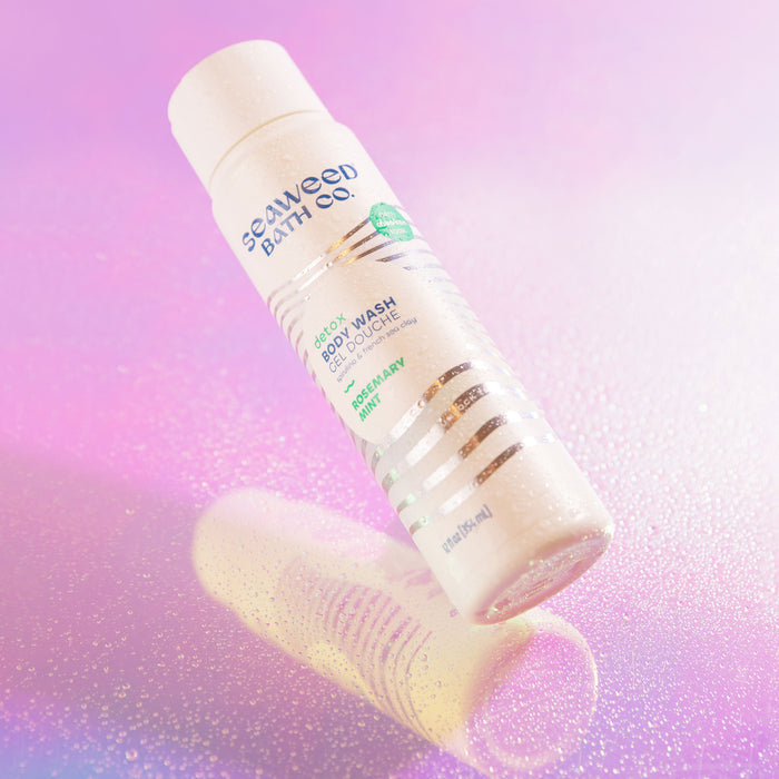 A bottle of The Seaweed Bath Cos Detox Purifying Body Wash, enriched with eucalyptus, peppermint, and Bladderwrack Seaweed for a refreshing cleanse, is featured against a pink and purple gradient background. Its slightly tilted surface is adorned with droplets.