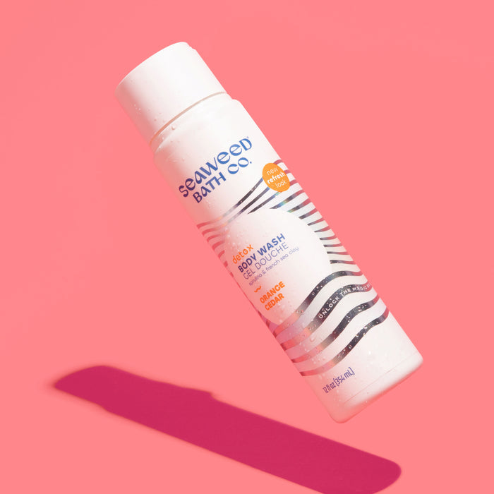 A 12 fl. oz. bottle of The Seaweed Bath Co.s Detox Purifying Body Wash in Awaken Ginger Cedar scent appears against a pink background, its white exterior adorned with black stripes and orange accents, infused with Green Coffee Bean Extract and Bladderwrack Seaweed.