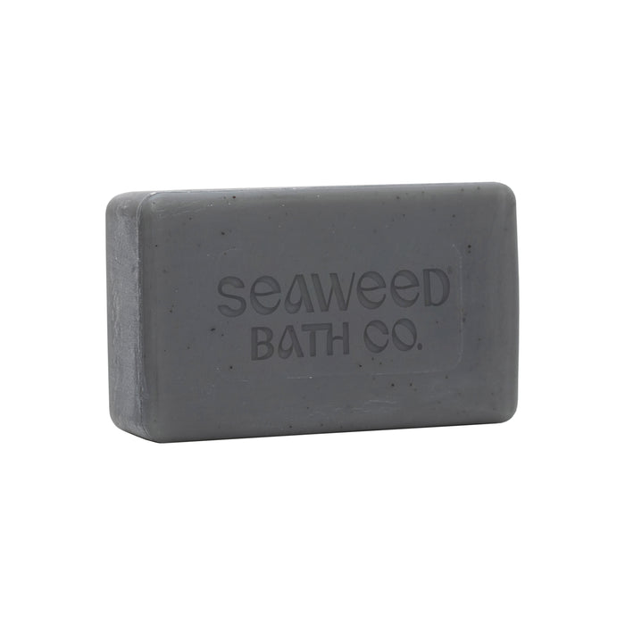 A rectangular gray Detox Purifying Facial Bar by The Seaweed Bath Co. is embossed on its surface and infused with detox properties, featuring bladderwrack seaweed extract and charcoal powder for a smooth texture with fine speckles, offering a revitalizing cleanse.