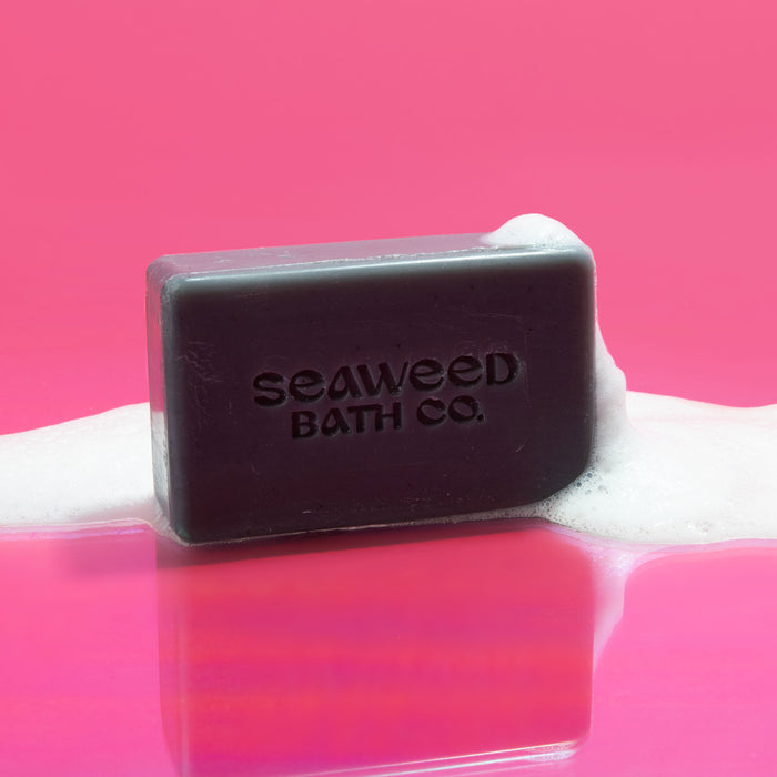 The Detox Purifying Facial Bar by The Seaweed Bath Co rests on a pink surface, surrounded by white foam. Featuring bladderwrack seaweed extract, the facial bar is complemented by the vibrant pink background reflecting off the wet surface.