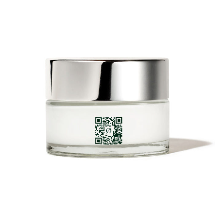 A small, clear glass jar with a silver lid contains IZZY Zero Waste Beautys Dew Bomb Glowy Hydra-Milk Starter Kit, a white cream with hydrating milk and hyaluronic acid. A green QR code on its side casts a shadow beneath, set against a plain white background.