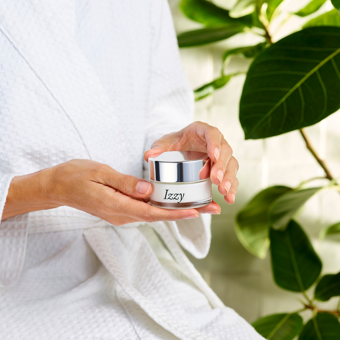 A person in a white bathrobe holds an IZZY Zero Waste Beauty Dew Bomb Glowy Hydra-Milk Starter Kit jar with a silver lid, surrounded by large green leaves and bathed in soft natural light.