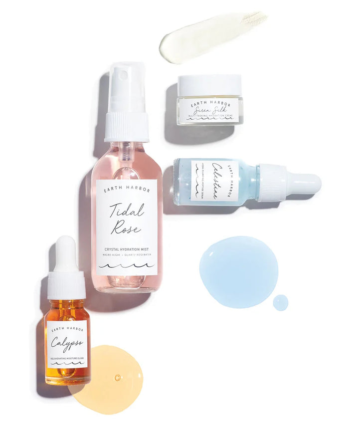 A flat lay of Earth Harbors DEWY YOU Kit on a white background, featuring a pink spray bottle, two jars with cream and gel, a blue hydrating skin serum dropper, an orange oil dropper, and product smears.