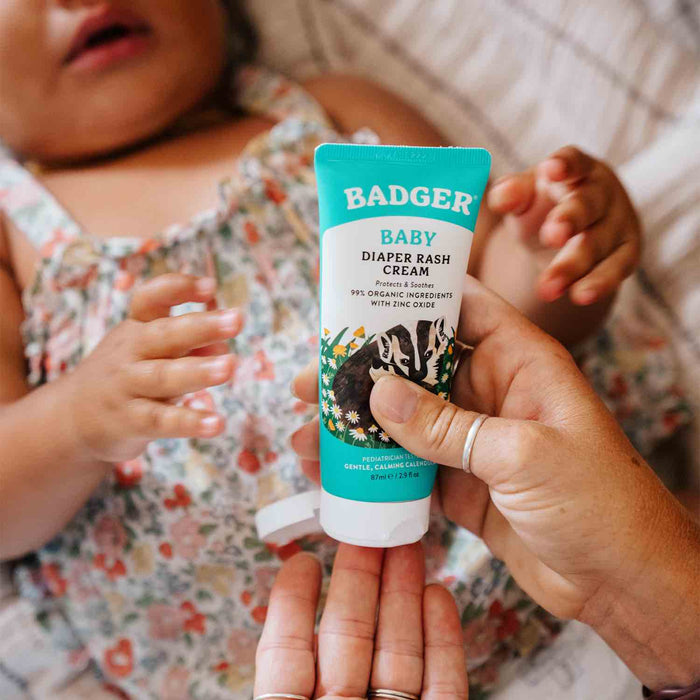 Someone holds a tube of Badger Balm Diaper Cream with a green and white label featuring non-nano zinc oxide. In the background, a baby in a floral outfit reaches for the cream on a patterned bedspread.