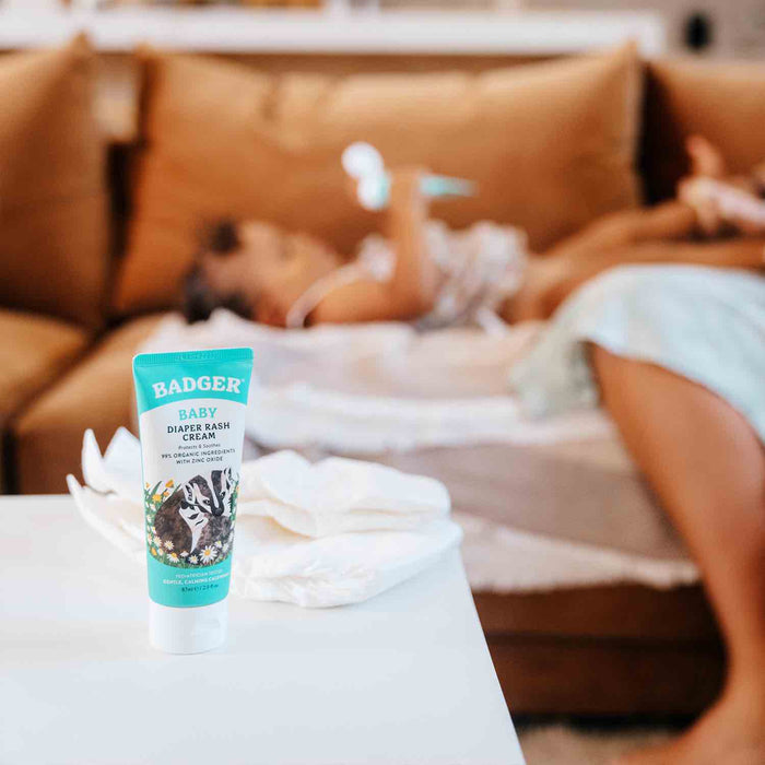 A tube of Badger Balm Diaper Cream with organic calendula is on a white table. In the background, a baby is being changed on a softly lit brown couch surrounded by scattered diapers. The cream contains non-nano zinc oxide for gentle relief.
