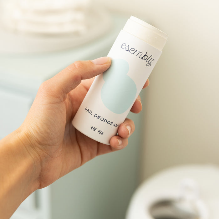 A person holds a white cylindrical container of Esembly Baby Diaper Pail Odor Eliminator & Deodorizer, showcasing a blue abstract design. This all-natural powder subtly brightens the softly lit scene, while the blurred background hints at a light-colored bathroom setting.