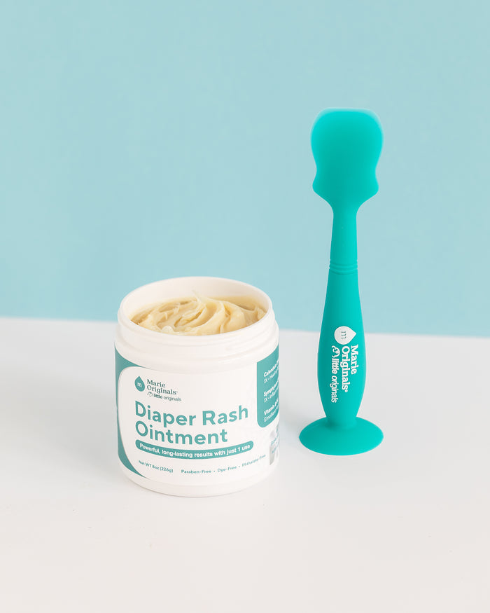 A jar of diaper rash ointment with a teal label rests beside a Marie Originals Diaper Spatula, which has a handy suction cup base. The background features a gradient of light blue and white.