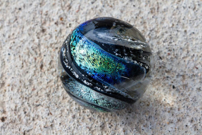 Round dichroic glass cremains marble with black edge and rainbow and blue gold copper swirls encasing ashes that will roll.