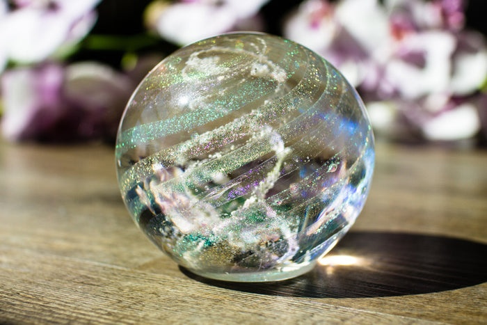 Dichroic rainbow glass orb containing cremation ashes with glittery ribbons, resting on a wooden surface.