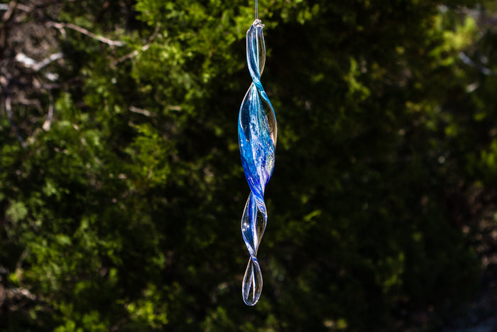 Dichroic spinner suncatcher infused with cremains with tapered ends and a wide center hanging in sunlight.