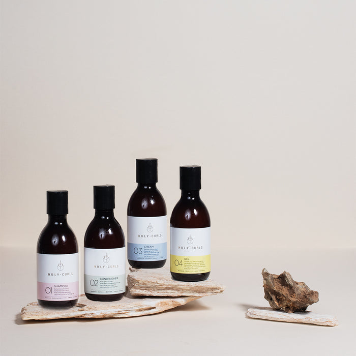 The Holy Curls Discovery Kit, featuring four bottles labeled 01 Shampoo, 02 Conditioner, 03 Cream, and 04 Gel, is elegantly displayed on wooden pieces against a neutral background. A small rock nearby enhances the minimalist aesthetic of this perfect curl routine.