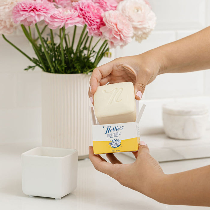 Someone holds a Nellies Dish Butter Refill outside its yellow and white box, reflecting eco-friendly cleaning. A vase of pink and white flowers is seen beside a white container on the countertop, advocating for a zero-waste lifestyle.