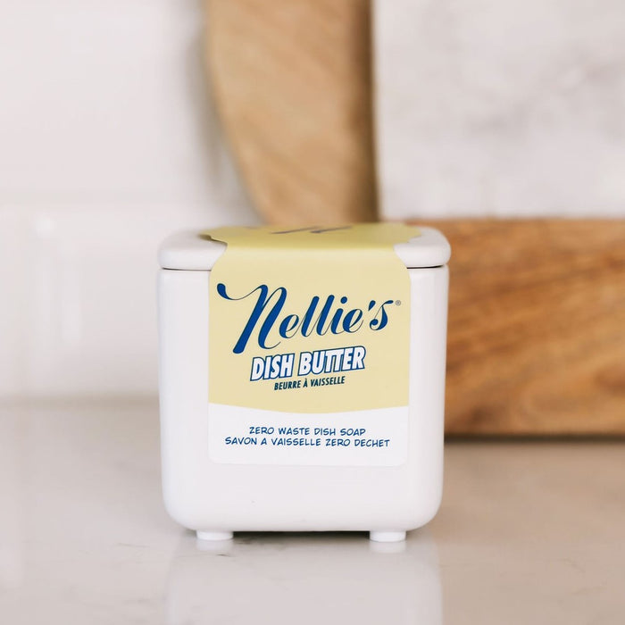 A white container of Nellies Dish Butter with a yellow label sits on the countertop, promoting eco-friendly cleaning with Zero Waste Dish Soap in English and French. A wooden cutting board is visible in the background.