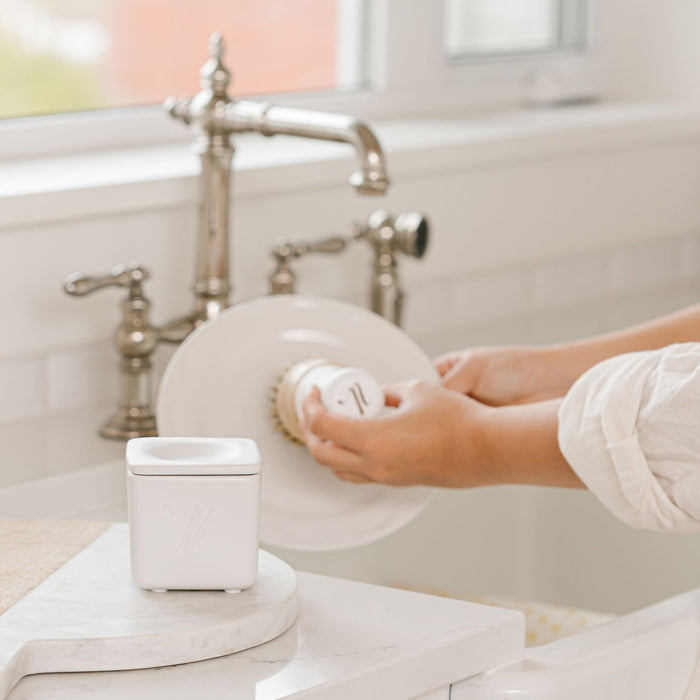 A person scrubs a white plate using Nellies Dish Butter with a soap brush at the kitchen sink. The metallic faucet gleams, and a white dispenser holds eco-friendly Dish Butter, making the bright, pristine space an epitome of zero-waste, green cleaning in this modern kitchen.