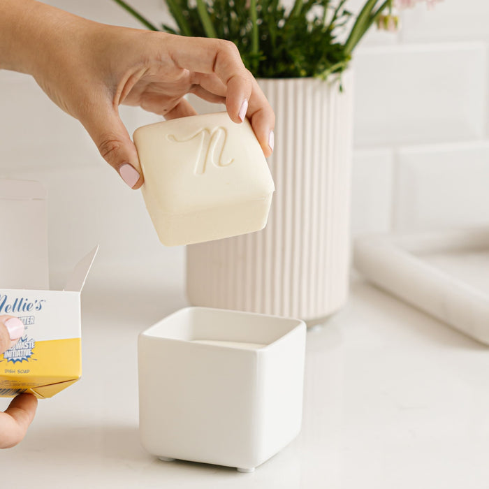 A hand places Nellies Dish Butter, a white, square soap with an n, into a white holder, reflecting zero-waste ideals. In the background, green-stemmed vases grace the countertop as the packaging subtly appears from the left.