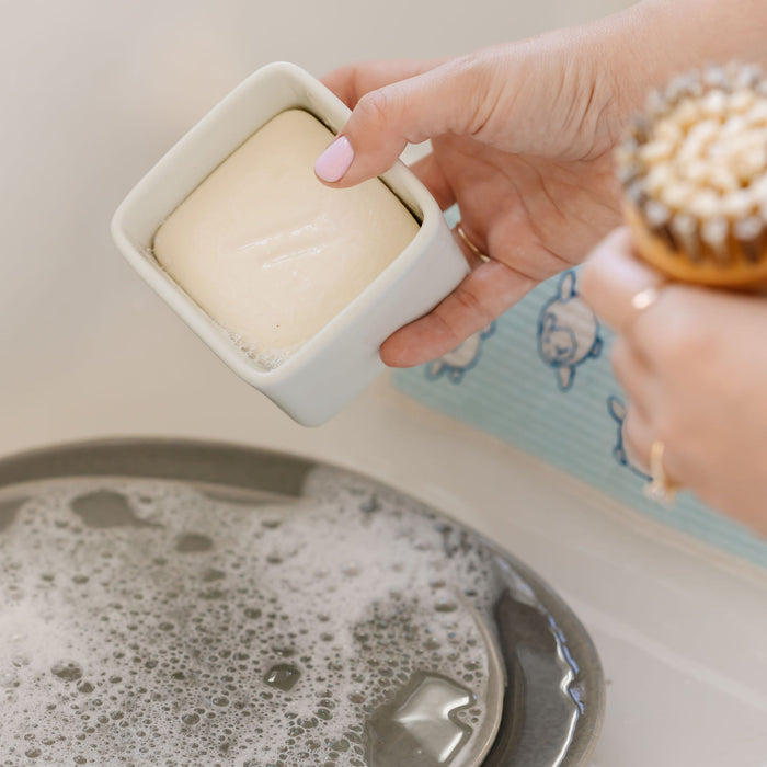 A person delicately holds Nellies Dish Butter with a soft brush over a basin of soapy water, embodying green cleaning. Light pink nail polish and rings add charm, while a sheep-adorned blue towel in the background enhances the sustainable elegance.
