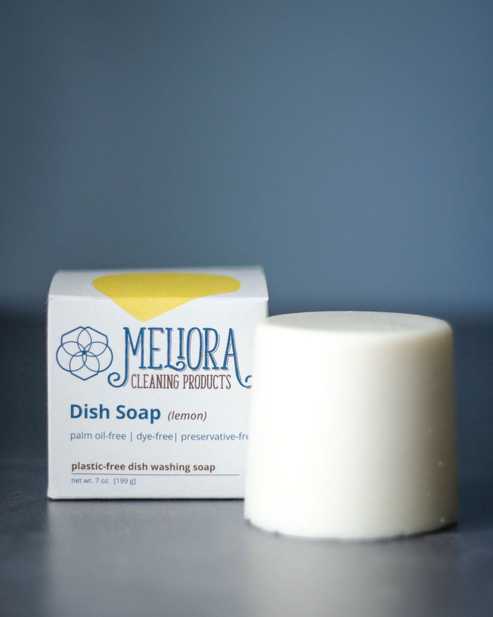 The Meliora Dish Soap Bar, with a refreshing lemon scent, is showcased alongside its white, blue, and yellow accented box. This zero-waste soap is plastic-free and contains no palm oil, dyes, or preservatives.
