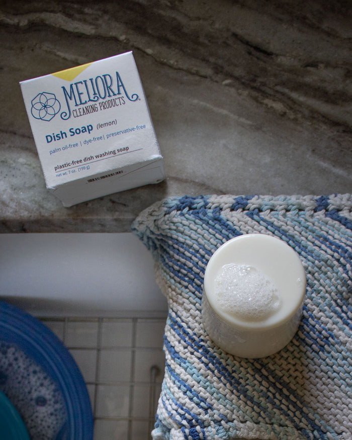 A box containing Melioras Dish Soap Bar, recognized for its zero-waste, plastic-free packaging, rests on a textured blue and beige cloth. Nearby is a white container with soapy bubbles, accompanied by glimpses of blue dishes and a sink basin in the background.