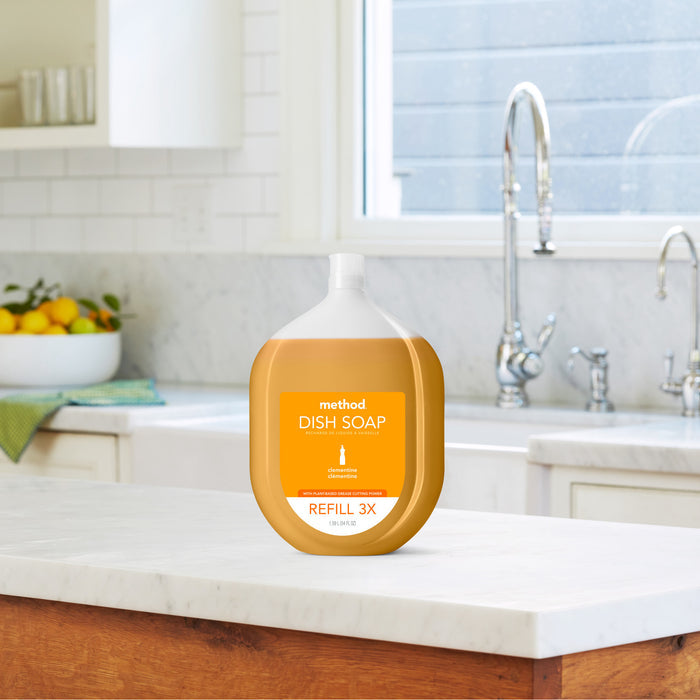A large pouch of methods Dish Soap Refill, known for its ultra grease-fighting power, stands on a kitchen counter. The eco-friendly kitchen, with recycled plastic decor, includes a marble countertop and bright window. A bowl of lemons and a towel add charm to the scene.