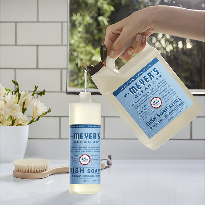 Someone refills a smaller bottle with Mrs. Meyers Dish Soap Refill in the kitchen. Natural cleaning agents release a fresh scent, as white flowers and a brush adorn the counter near a window framed by tiled walls in the background.